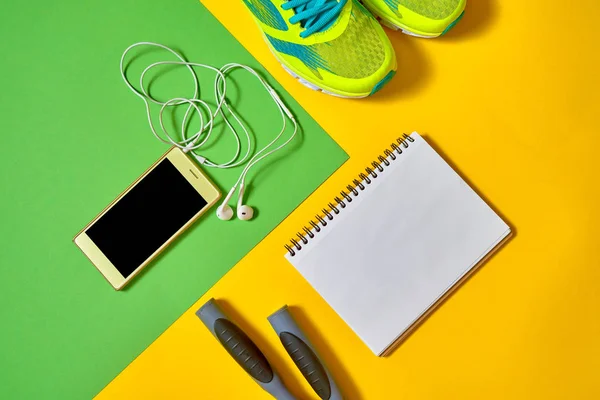 Sports equipment with shoes, skipping rope, blank notebook and mobile cellphone with earphones on colorful background. Top view, flat lay. Sport, fitness concept, healthy lifestyle