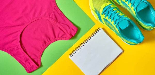 Sport shoes, woman\'s top bra and white blank notebook on colorful yellow and green backgroundon, copy space. Top view, flat lay. Sport and healthy lifestyle concept, flat lay