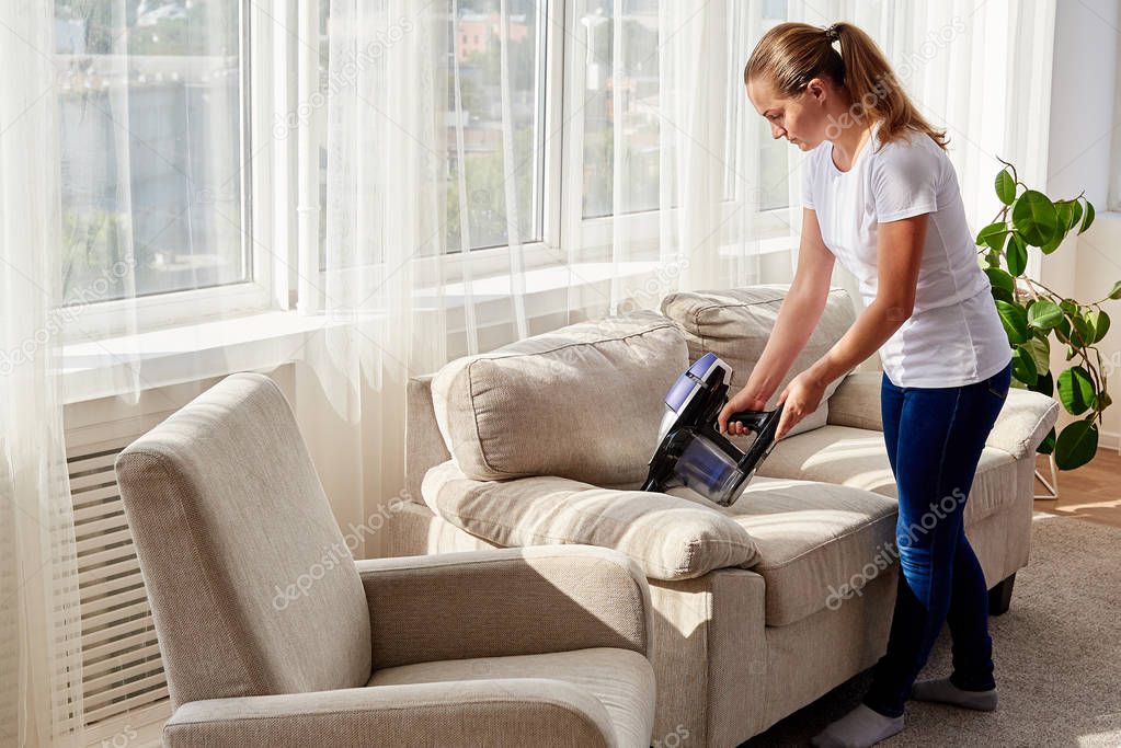 Full body portrait of young woman in white shirt and jeans cleaning sofa with vacuum cleaner in living room, copy space. Housework, cleanig and chores concept. House hygiene, cleaning vacuum appliance