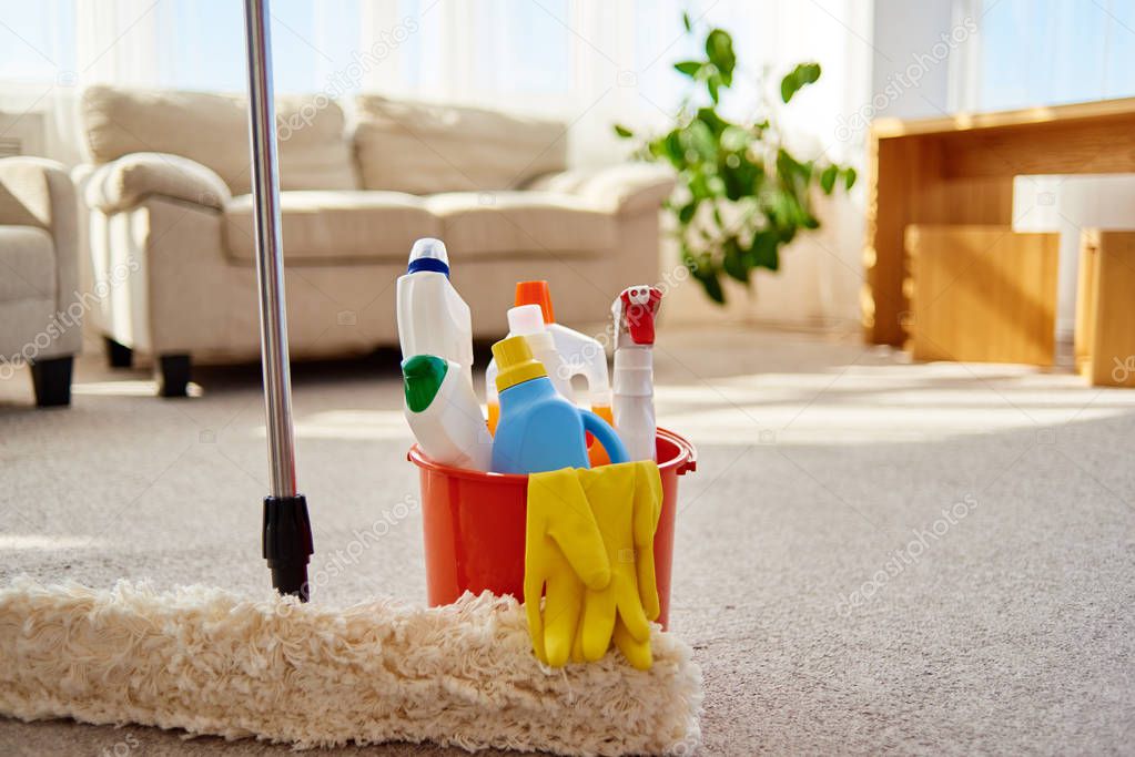 Cleaning set for different surfaces in orange bucket and mop on floor in living room, copy space. Cleaning service concept. Bucket with cleaning items for kitchen, bathroom or toilet