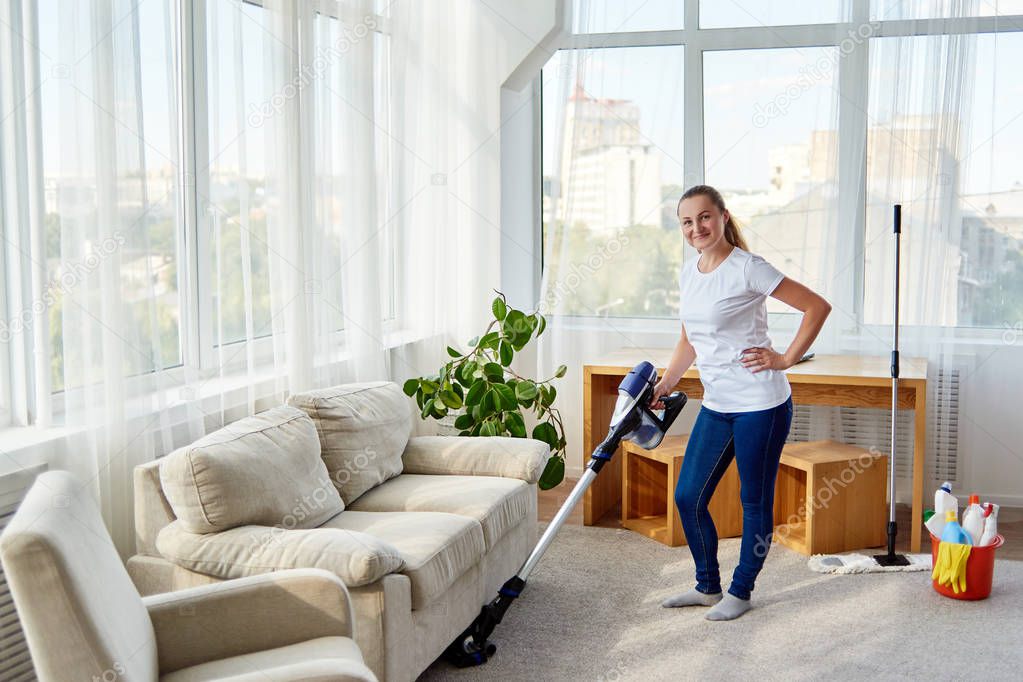 Full body portrait of smiling woman in white shirt and jeans cleaning carpet with vacuum cleaner in living room, copy space. Housework, cleanig and chores concept