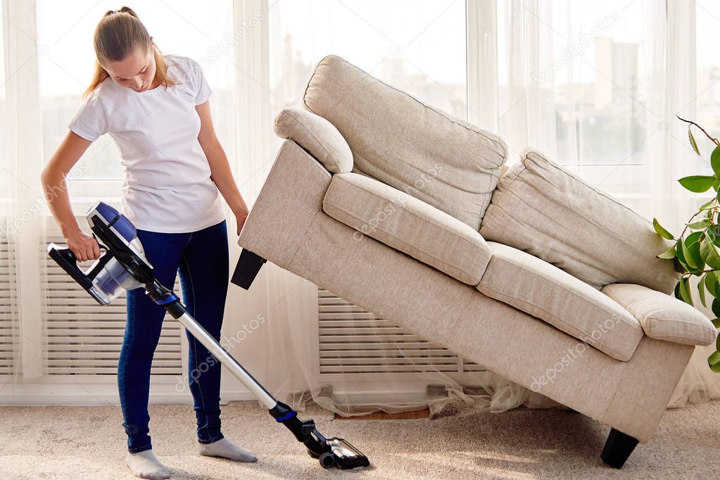 Full length body portrait of young woman in white shirt and jeans cleaning carpet with vacuum cleaner under sofa in living room, copy space. Housework, cleanig and chores concept