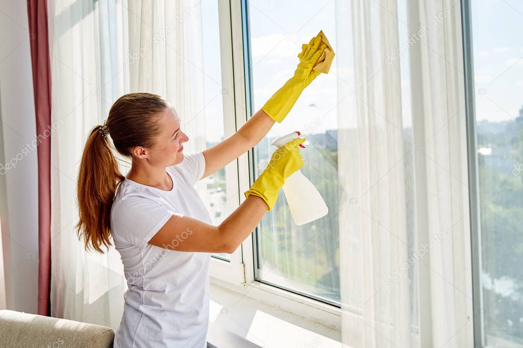 Woman in white shirt and yellow rubber gloves cleaning window with cleanser spray and yellow rag at home or office, copy space, back view. People, housework and housekeeping concept
