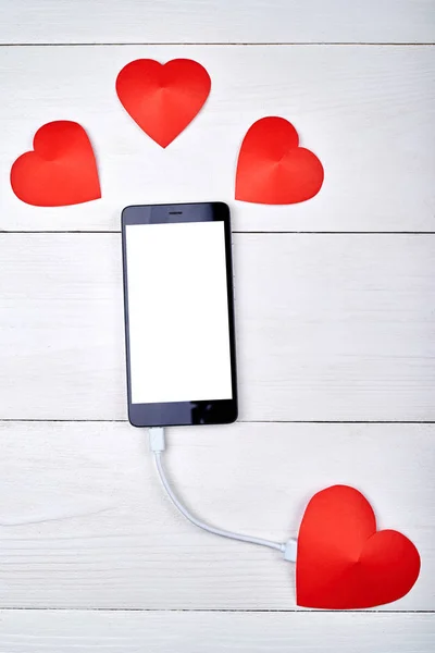 Mobile smart phone with white blank screen charging with red paper heart on white wooden background, copy space. Mockup template for Valentines Day. Love, technology concept. Top view, flat lay
