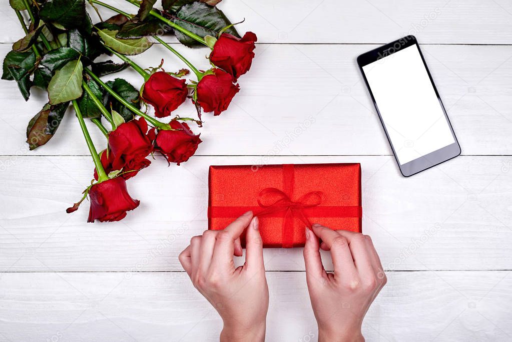 Woman wrapping red gift box with bouquet of roses and mobile cellphone with blank screen on white background, copy space. Flat lay, top view. Valentines day, Womens Day, Mothers Day