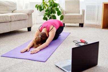 Güzel genç bir kadın evde yerde esneme egzersizi yapıyor, dizüstü bilgisayarda online eğitim alıyor, kopyalama alanı kullanıyor. Tam boy portre. Yoga, pilates, egzersiz.