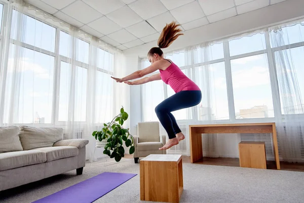 Young woman doing box jump exercise in living room at home, copy space. Jumping squats. Sport, healthy lifestyle concept
