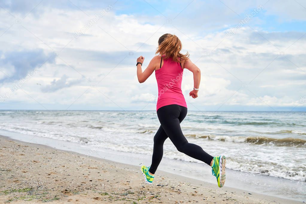 Back view of young fitness woman runner running at seaside, copy space. Girl working out on beach at summer morning, full length portrait. Healthy lifestyle concept