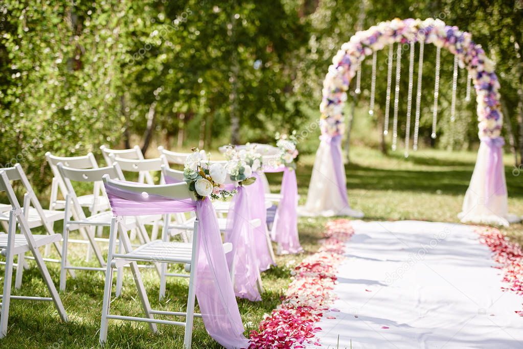 Place for wedding ceremony. Wedding arch decorated with cloth and flowers and chairs on each side of archway outdoors, copy space. Empty wooden chairs for guests on green grass. Wedding setup