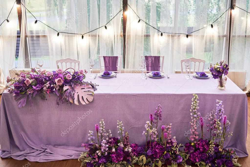 Wedding presidium in restaurant, copy space. Banquet table for newlyweds with flowers, monstera leaves, violet cloth and bulbs. Lush floral arrangement. Luxury wedding decorations 