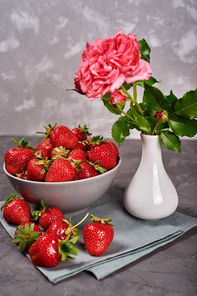 Red ripe strawberries in ceramic bowl on linen table napkin and pink rose in vase on gray concrete background, copy space. Healthy food concept, still life