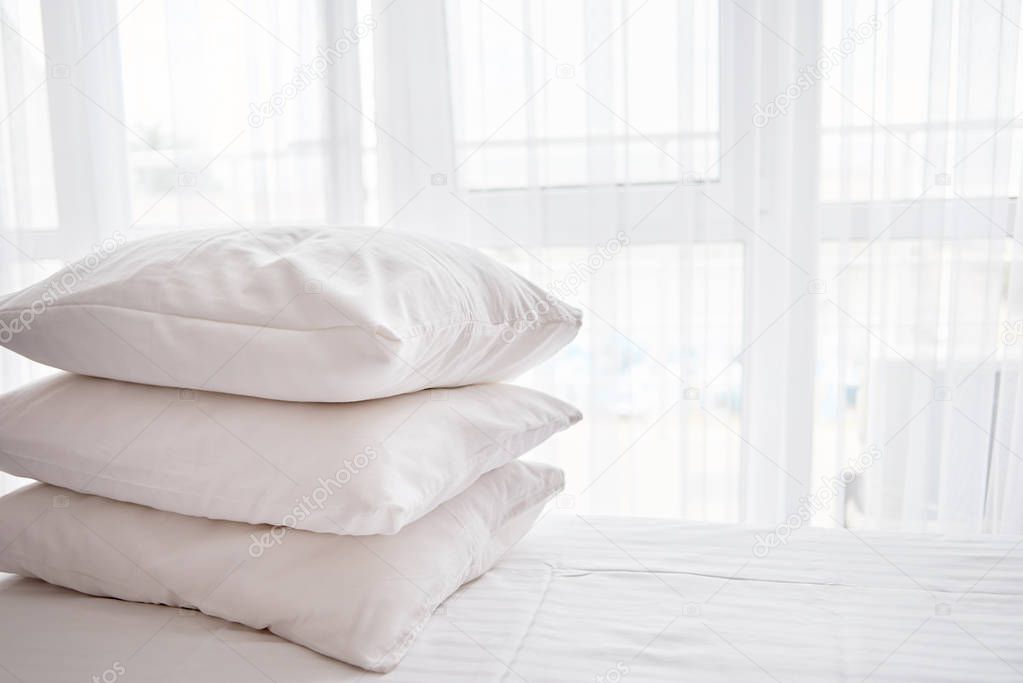 Stack of white soft pillows on comfortable bed sheet with window on background, copy space
