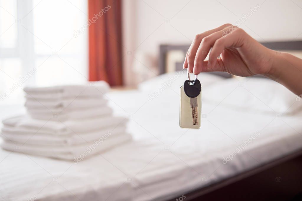Woman holding in hand room key with blank label at hotel suite, copy space. Bedroom interior in blur