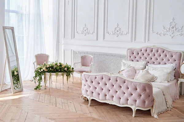 Big comfortable pink royal bed with pillows in elegant bedroom interior, copy space. Honeymoon suite with mirror and flowers on table. Luxury bed in romantic style. Boudoir