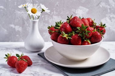 Keten masa peçete ve gri alçı arka plan üzerinde vazo papatya çiçekleri üzerinde beyaz seramik kase kırmızı olgun çilek, kopya alanı. Sağlıklı gıda kavramı, natürmort