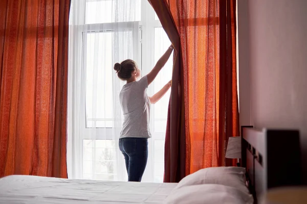 Beautiful young woman standing in hotel room, opening curtains and looking through the window