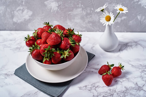 Red ripe strawberries in white ceramic bowl on linen table napkin and chamomile flowers in vase on grey plaster background, copy space. Healthy food concept, still life