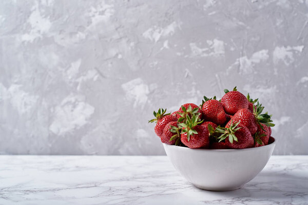 Organic ripe strawberries in white ceramic bowl on gray plaster wall background, copy space. Healthy food concept, still life