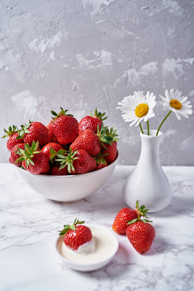 Red organic strawberries in white ceramic bowl, cream and chamomile flowers in vase on gray marble background, copy space. Healthy food concept, still life