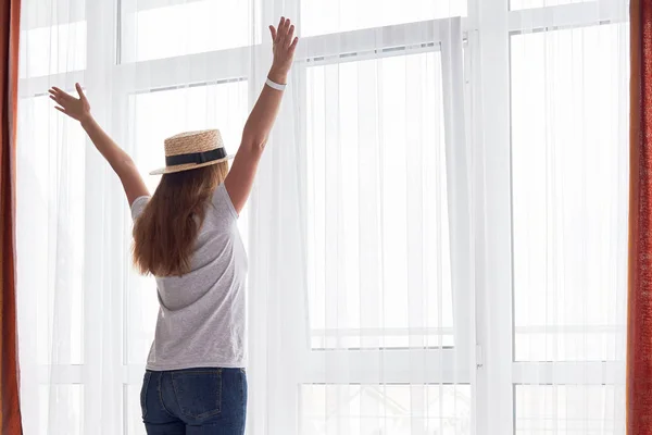 Happy young woman in hat staying with raised up arms in hotel room near big french window, copy space