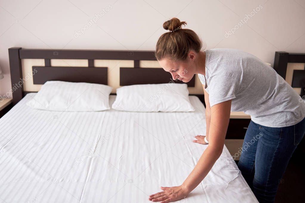 Beautiful young woman making bed at home or in hotel room, copy 