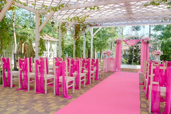 Place for wedding ceremony with wedding arch decorated with flowers and pink cloth outdoors, copy space. Wedding decorations with chairs