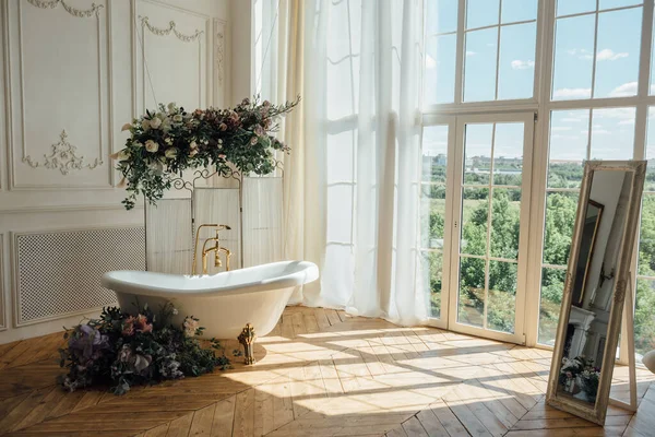 Beautiful luxury vintage empty bathtub with lush floral decorations and candles, mirror on the floor in bathroom interio, copy space. Freestanding white bath with flowers near folding screen