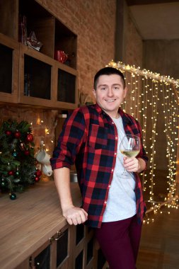 Happy smiling man holding glass of white wine in kitchen at home with Christmas decor and garland on background, copy space