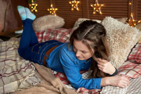 穿着牛仔裤的漂亮女人躺在床上 卧室的木板墙上挂着花环 复制空间 舒适的家的时刻 圣诞节的概念 — 图库照片