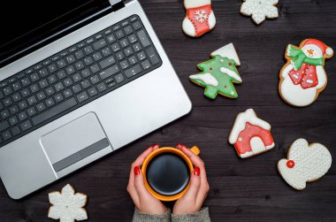 Karanlık masada elinde bir fincan sıcak kahve tutan bir kadın dizüstü bilgisayarlı ve Noel kurabiyeli kurabiye, fotokopi odası. Üst manzara, düz yatış. Tatil kavramı