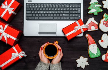 Karanlık masada elinde bir fincan sıcak kahve, açık dizüstü bilgisayar, kırmızı hediye kutuları ve Noel kurabiyeleri olan bir kadın, fotokopi odası. Üst manzara, düz yatış. Tatil kavramı