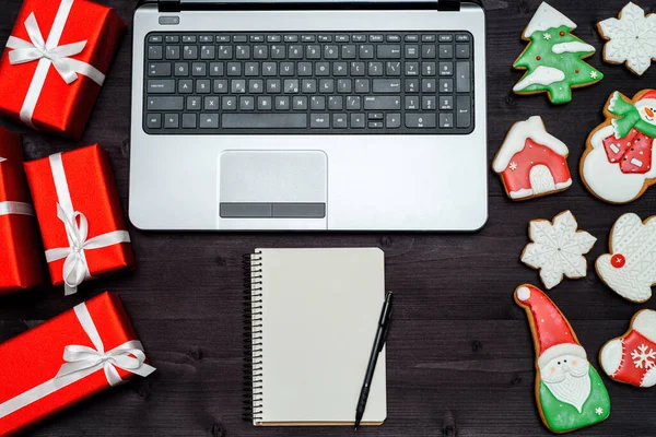 Open laptop computer, Christmas presents, blank notebook and gingerbread cookies on dark wood background, copy space. Top view, flat lay. Santa, snowflake, mitten, house, stocking, snowman, fir tree