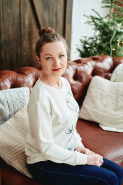 Beyaz kazaklı ve kot pantolonlu bir kadın arka planda köknar ağacı olan kahverengi deri koltukta oturuyor. Noel arşivi 