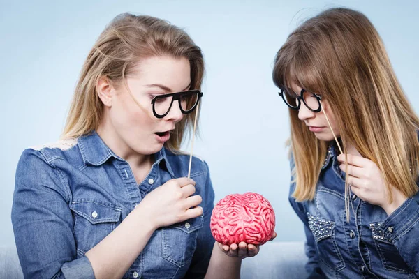 Friendship, human relations concept. Two crazy women friends or sisters wearing jeans shirts and eyeglasses on stick, thinking about solving problem holding fake brain