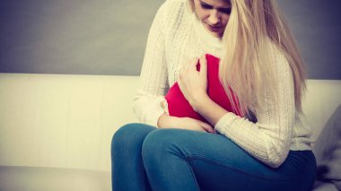 Acı dolu dönemler ve adet krampı sorunları konsepti. Karnı ağrıyan bir kadın kahve içerken kendini kötü hisseder sıcak su şişesini tutar ve biraz rahatlar.