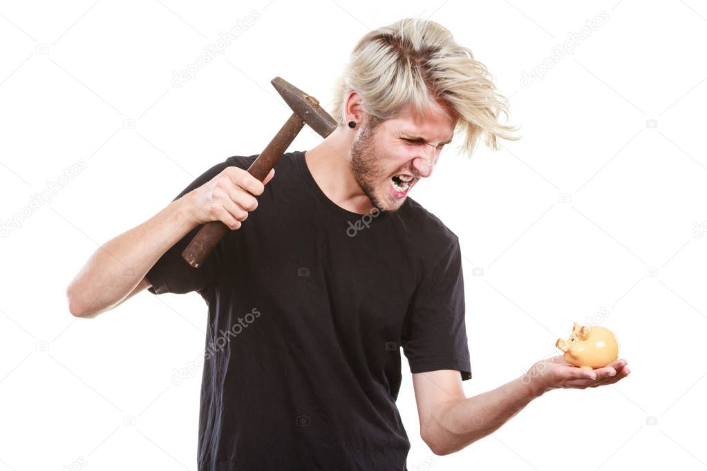 Money, savings, finances concept. Angry sreaming young blonde man wearing black t shirt trying to break piggy bank with hammer, studio shot isolated