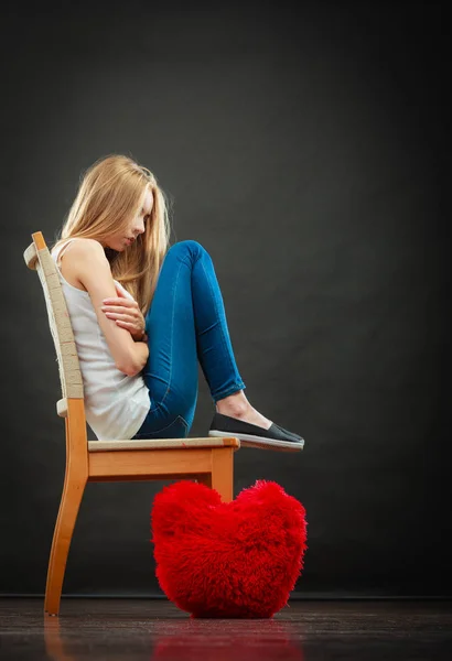 Broken heart love concept. Sad unhappy woman sitting on chair red heart pillow on floor dark background