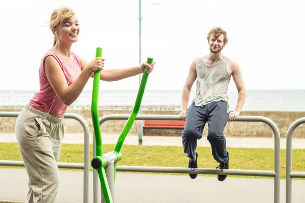 Active woman exercising on elliptical trainer machine and man listening to music. Happy fit sporty girl in training suit working out at outdoor gym. Sport fitness and healthy lifestyle concept.