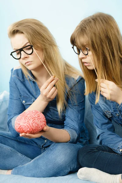 Two serious women friends or sisters wearing jeans shirts and eyeglasses on stick, thinking about solving problem holding fake brain