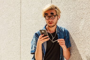 Erkek moda, teknoloji, kentsel tarzı giyim kavramı. Müzik ve telefon tutarak şehir sokak giyen kot kıyafeti ve eksantrik güneş gözlüğü üzerinde duran hipster adam