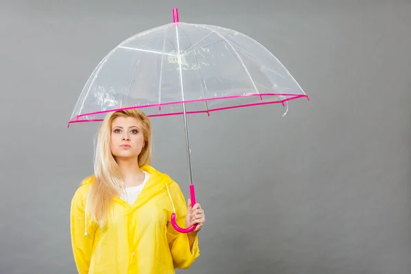 Sad bored blonde woman wearing yellow raincoat holding transparent umbrella waiting for rain.