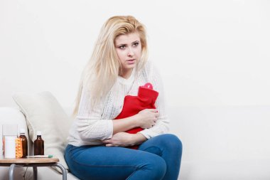 Acı dolu dönemler ve adet krampı sorunları konsepti. Karnı ağrıyan bir kadın kahve içerken kendini kötü hisseder sıcak su şişesini tutar ve biraz rahatlar.