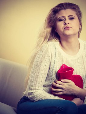 Acı dolu dönemler ve adet krampı sorunları konsepti. Karnı ağrıyan bir kadın kahve içerken kendini kötü hisseder sıcak su şişesini tutar ve biraz rahatlar.