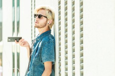 Erkek moda, teknoloji, kentsel tarzı giyim kavramı. Şehir sokak giyen kot kıyafeti ve kulaklık üzerinden müzik güneş gözlüğü üzerinde duran hipster adam