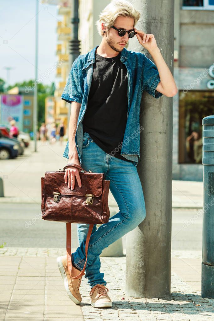 Men fashion, urban style clothing concept. Hipster guy walking through city wearing jeans outfit, male handbag and sunglasses on sunny day