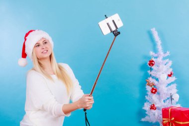 Noel hediyelerinin tadını çıkarıyorum. Noel Baba şapkalı bir kadın selfie çubuğu kullanarak kendi fotoğrafını çekiyor. Mavi arkaplanda kapalı çekim