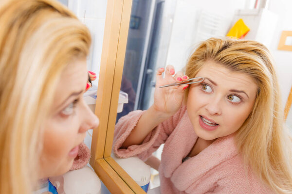 Woman in bathroom plucking eyebrows depilating with tweezers, looking at mirror. Girl tweezing removing her facial hairs. Wide angle view