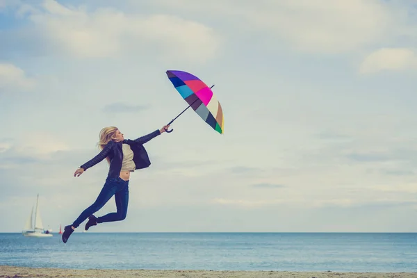 Happiness, enjoying cold autumn weather, feeling great concept. Woman jumping with colorful umbrella on beach near sea, sunny day and clear blue sky