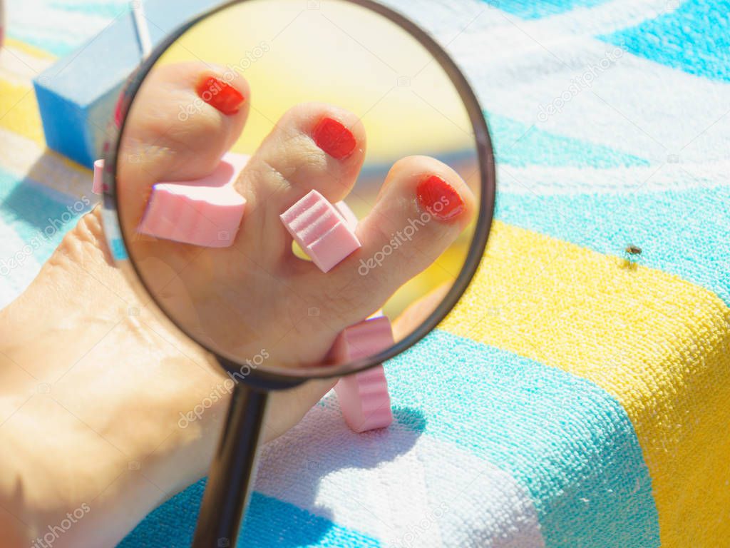 Woman looking at her fresh made pedicure through magnifying glass on towel. Female having toes nails painted with red nail polish.