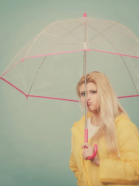 Sad bored blonde woman wearing yellow raincoat holding transparent umbrella waiting for rain.